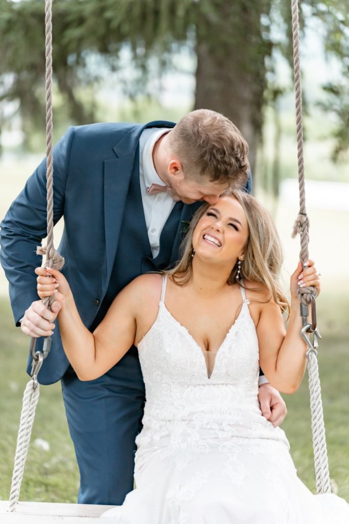 a groom kissing his bride on the forehead as she sits on a rustic swing